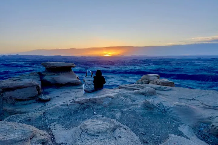 Zwei Personen sitzen am Rand einer Klippe und beobachten den Sonnenaufgang in der Wüste.